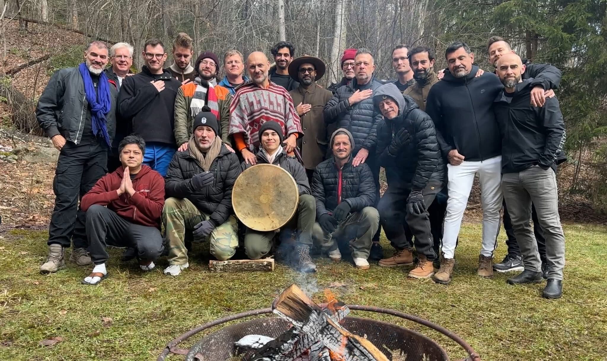 A gathering of men around a fire in winter woods, one holding a frame drum